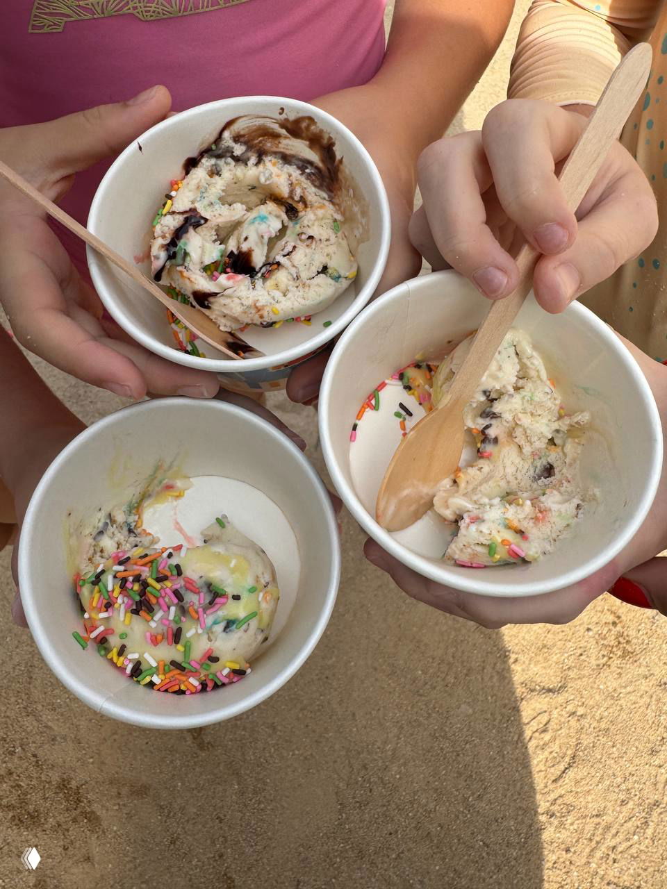 Три бумажных стаканчика с мороженым и разноцветной посыпкой, которые держат дети на песке — десерт для маленьких гостей клуба.