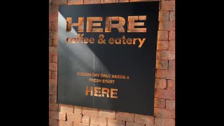 Вывеска кафе HERE coffee & eatery на кирпичной стене; чёрная табличка с крупными золотыми буквами и слоганом.