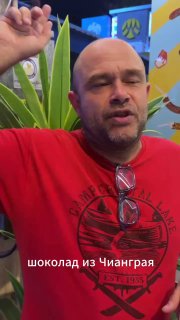 Портрет владельца ларька Dip Daddy, мужчина говорит что-то на камеру; субтитр на снимке: «шоколад из Чианграя», фон с промо‑артом ларька.