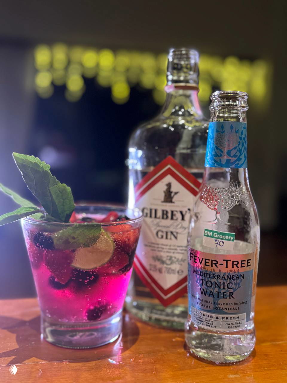 Яркий джин-тоник с замороженными ягодами, лаймом и веточкой мяты в стакане; на столе видна бутылка Gilbey's и медиттерранский тоник Fever-Tree.