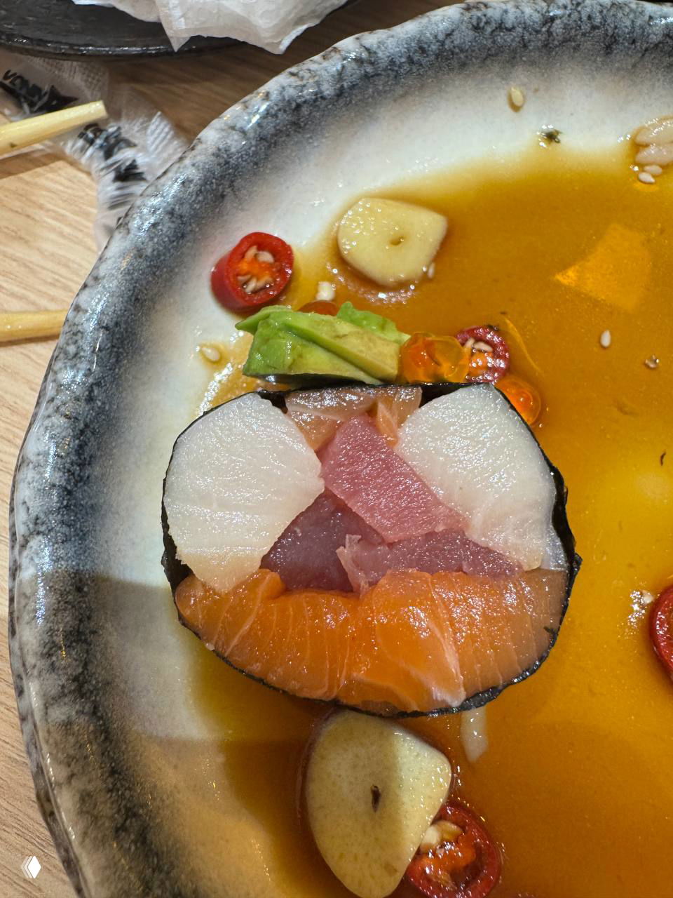 Поперечный срез Kaisen roll показывает три вида рыбы внутри — видны лосось, тунец и хамачи; рядом дольки чеснока и ломтики чили на тарелке с соусом.