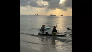 Два человека на сапах/каяках в мелкой воде у берега на фоне золотого неба и спокойного моря — активность у пляжа Taling Ngam.