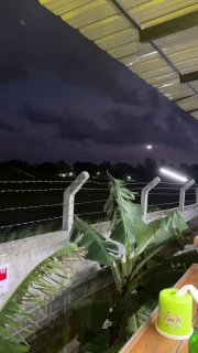 Вид с террасы через колючую проволоку на ночное небо и полосу; видна крыша навеса, освещение и растения у ограждения.