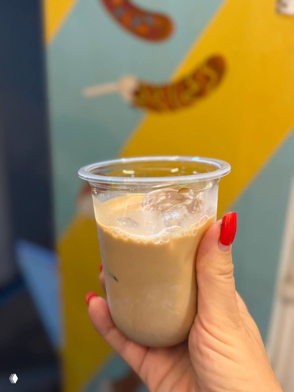 Пластиковый стакан с ледяным холодным напитком (cold brew/chai) в руке блогера, на заднем плане яркая стена с рисунками бананов и передний план в фокусе.