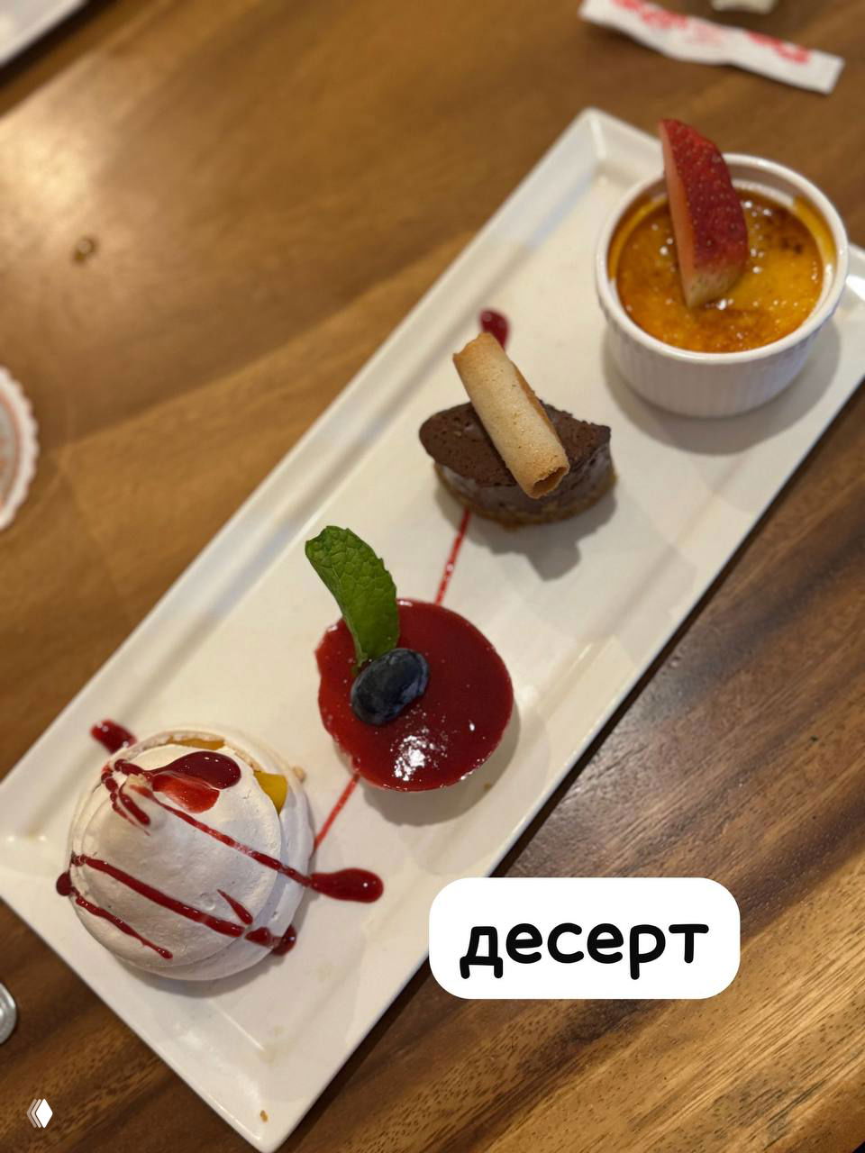 Квартет десертов на длинной белой тарелке: крем-брюле, шоколадный мини-десерт, мусс и Павлова — разнообразная финальная подача в сете.