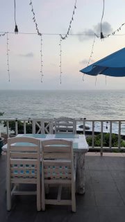 Терраса ресторана с белыми столами и стульями, зонтом и гирляндой — открытый вид на море и спокойный утренний берег острова Самуи.