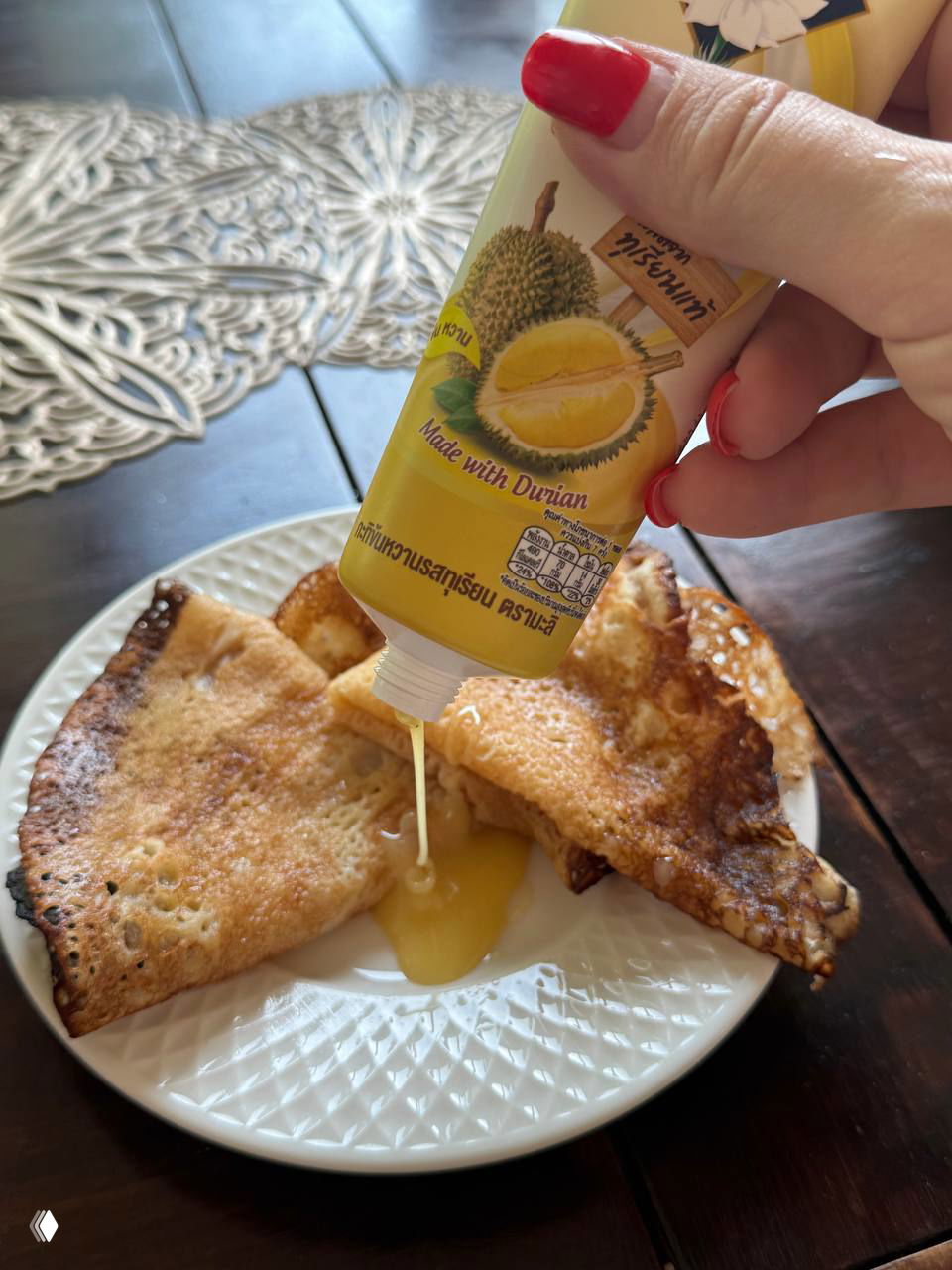 Рука держит тюбик дуриановой сгущёнки и выдавливает пасту на блины на тарелке — демонстрация продукта в употреблении.