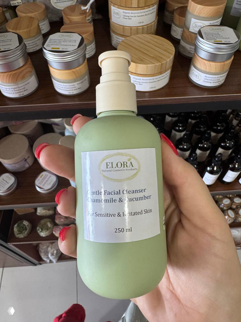Крупный план зелёной бутылки косметического средства ELORA Gentle Facial Cleanser (ромашка и огурец) на фоне полок с баночками и пробниками.