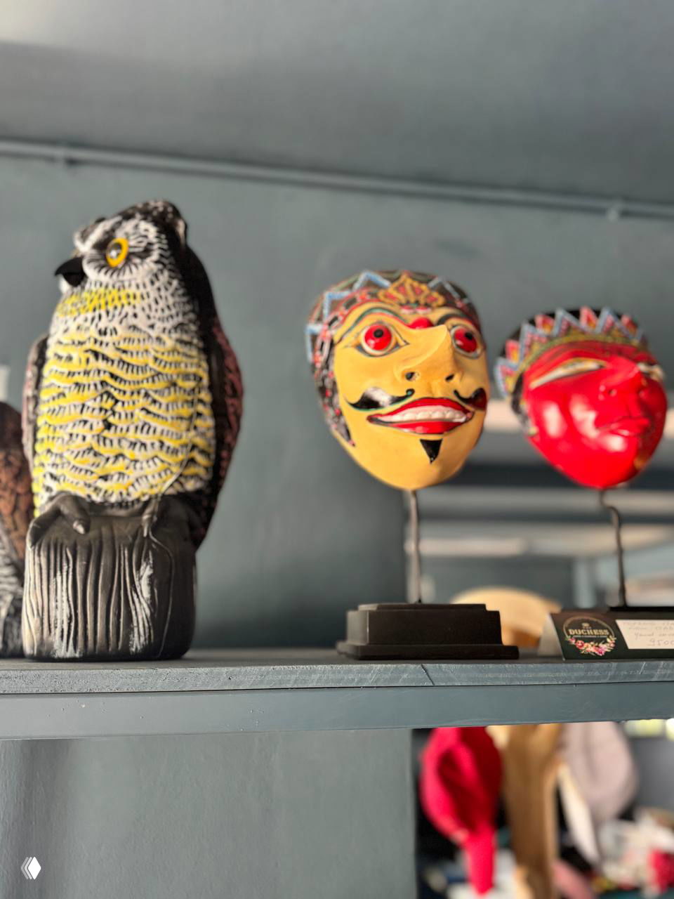 Керамическая фигурка совы и цветные маски на полке в магазине подарков: крупный план декоративных предметов на фоне полок и интерьера.