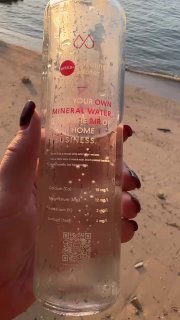 Рука держит прозрачную бутылку минеральной воды с заметной этикеткой и QR‑кодом на фоне пляжа и моря при закате рядом с Биг Буддой.