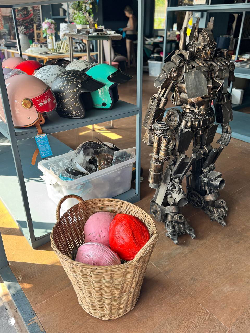 Скульптурная металлическая фигура-робот рядом с полкой, на которой лежат яркие шлемы и декоративные корзины — подборка необычных предметов для интерьера в магазине на Самуи.