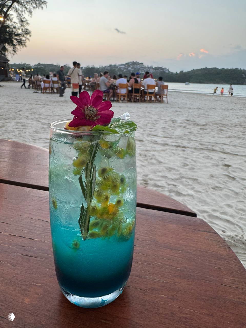 Голубой фирменный коктейль с цветком и ягодами на деревянном столе, на заднем плане виднеется пляж и столики Pi beach club.