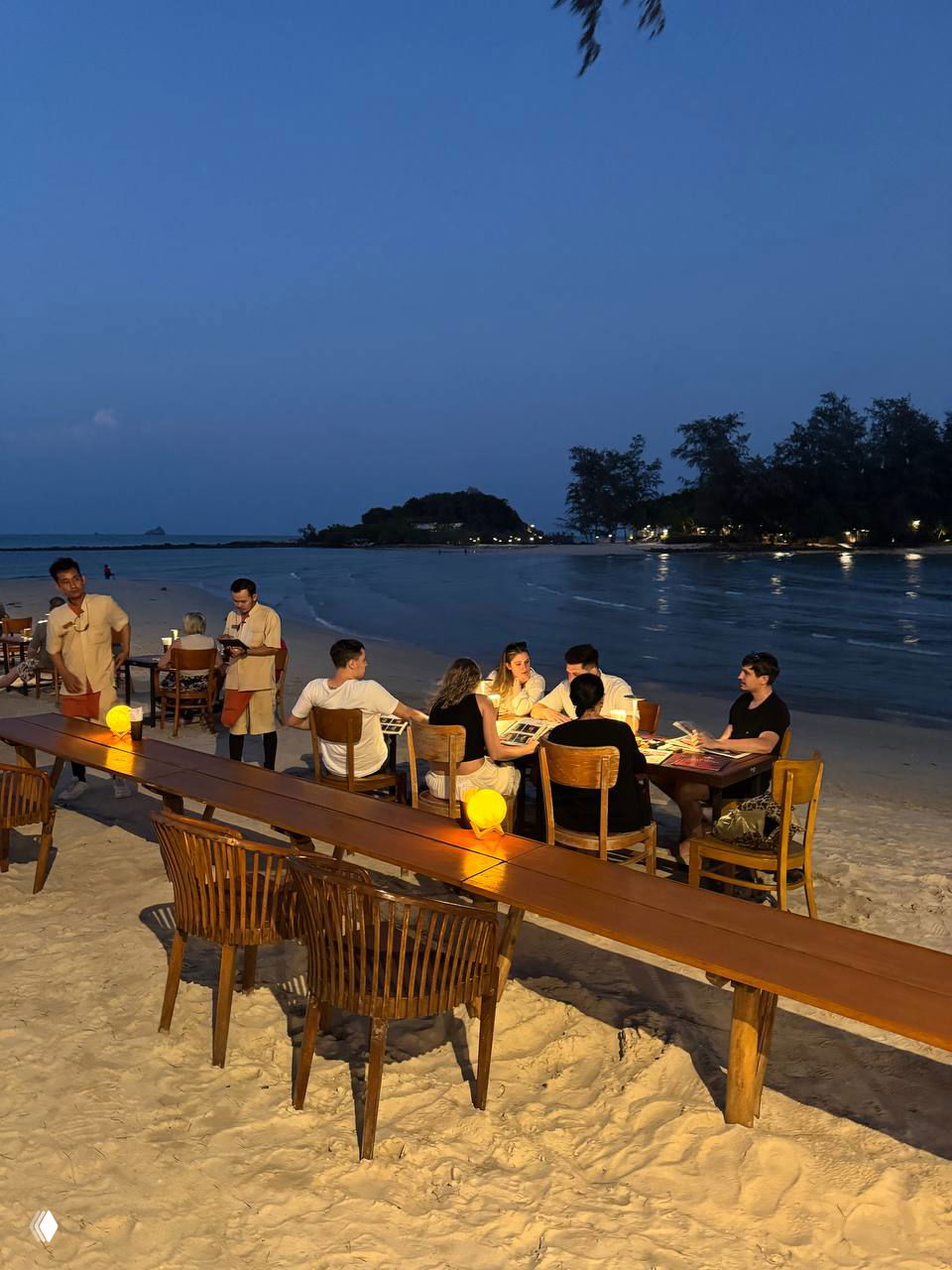 Вечерняя сервировка столиков на песке в Pi beach club, гости ужинают у моря при мягком освещении и спокойной прибрежной панораме.