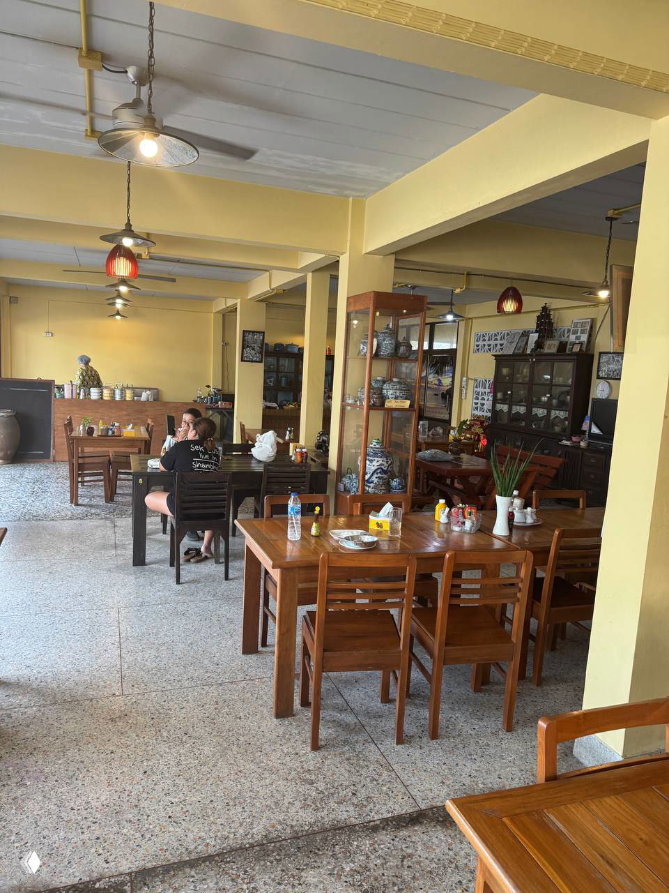 Интерьер кафе: деревянные столы и стулья в просторном зале, витрины с антиквариатом на фоне, естественное освещение и расслабленная обстановка.