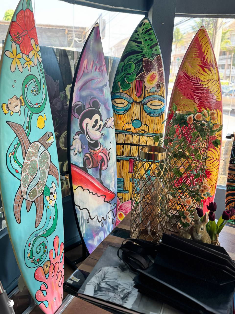 Декоративные серф-формы и расписанные доски с морскими мотивами на витрине — яркие настенные и напольные элементы для дома и кафе, представленные в магазине на Самуи.