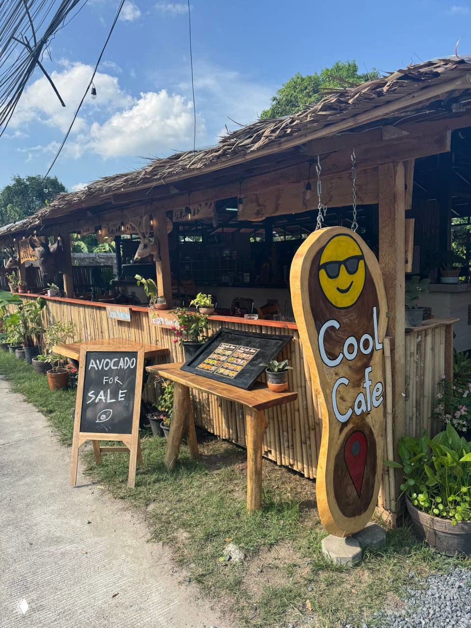 Фасад Cool Cafe в Ламае: деревянная вывеска с улыбающимся логотипом, меловая табличка «Avocado for sale» и стойка с меню у входа.