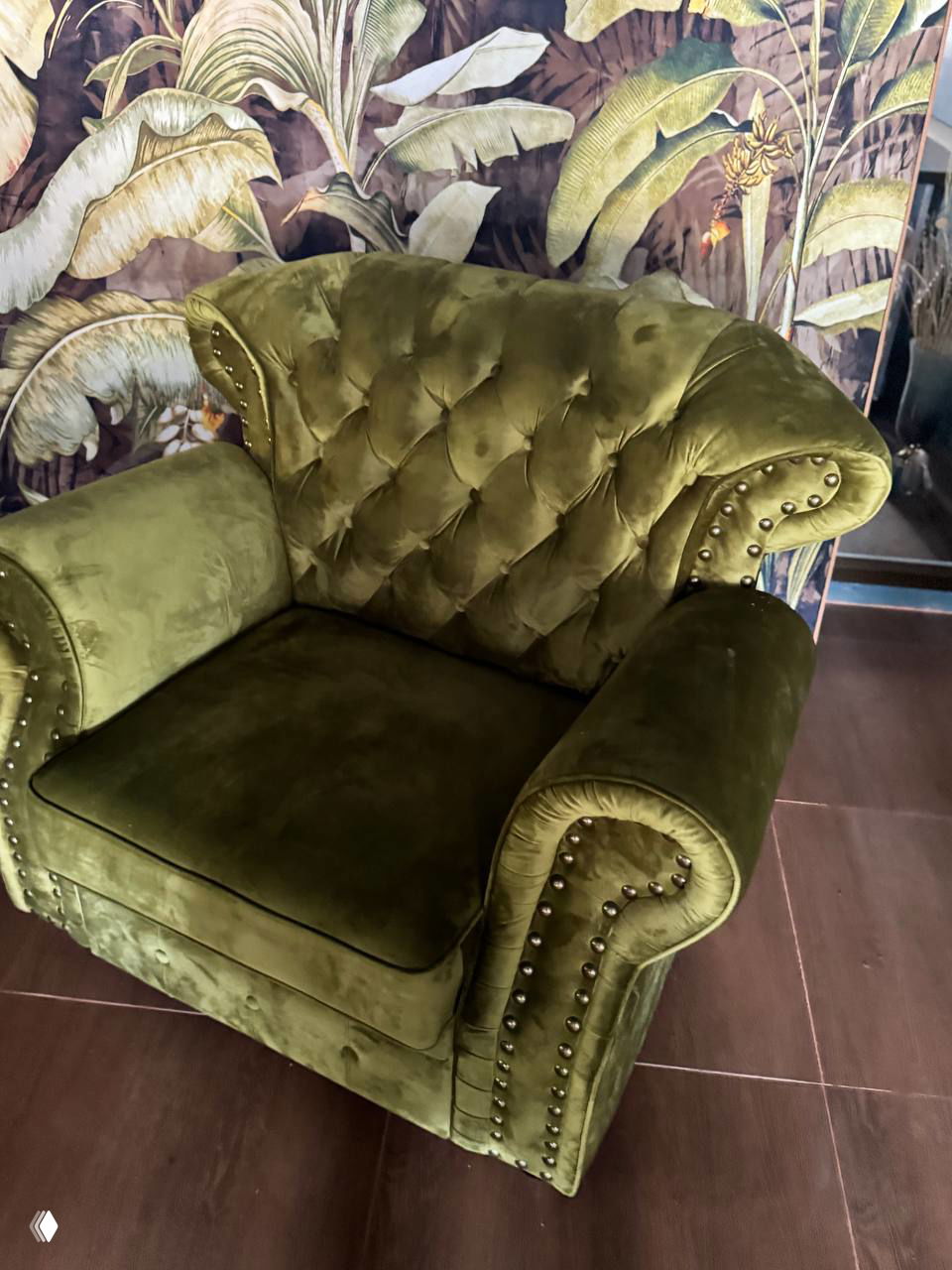 Зелёное бархатное кресло в винтажном стиле с мягкой обивкой и декоративной прострочкой, стоящее внутри магазина рядом с интерьерными аксессуарами.