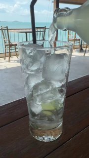 Стакан со льдом и долькой лайма, в который наливают прозрачный кокосовый цветочный сок на фоне моря и террасы кафе — подача напитка у моря на Самуи.