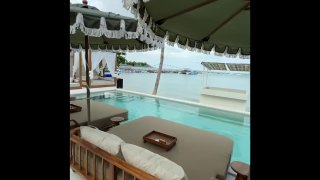 Зона отдыха у бассейна и моря в The Door beach club: новые диваны и шезлонги под зонтом, вид на причал и воду на заднем плане, атмосферный кадр у воды.