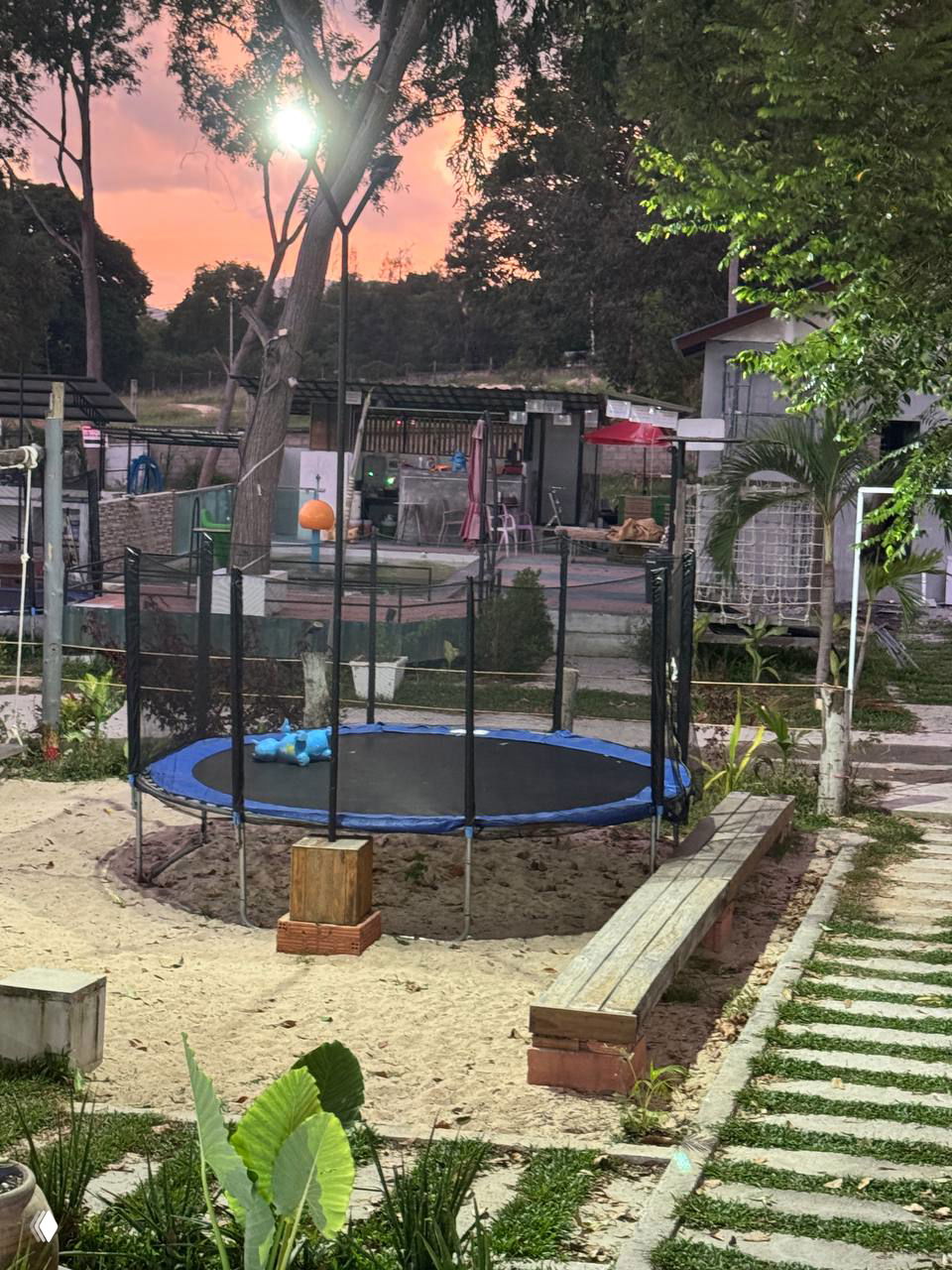 Зона у кафе с батутом и площадкой: батут в песке, лавочки и зелень, на заднем плане виднеется бассейн и служебные постройки.