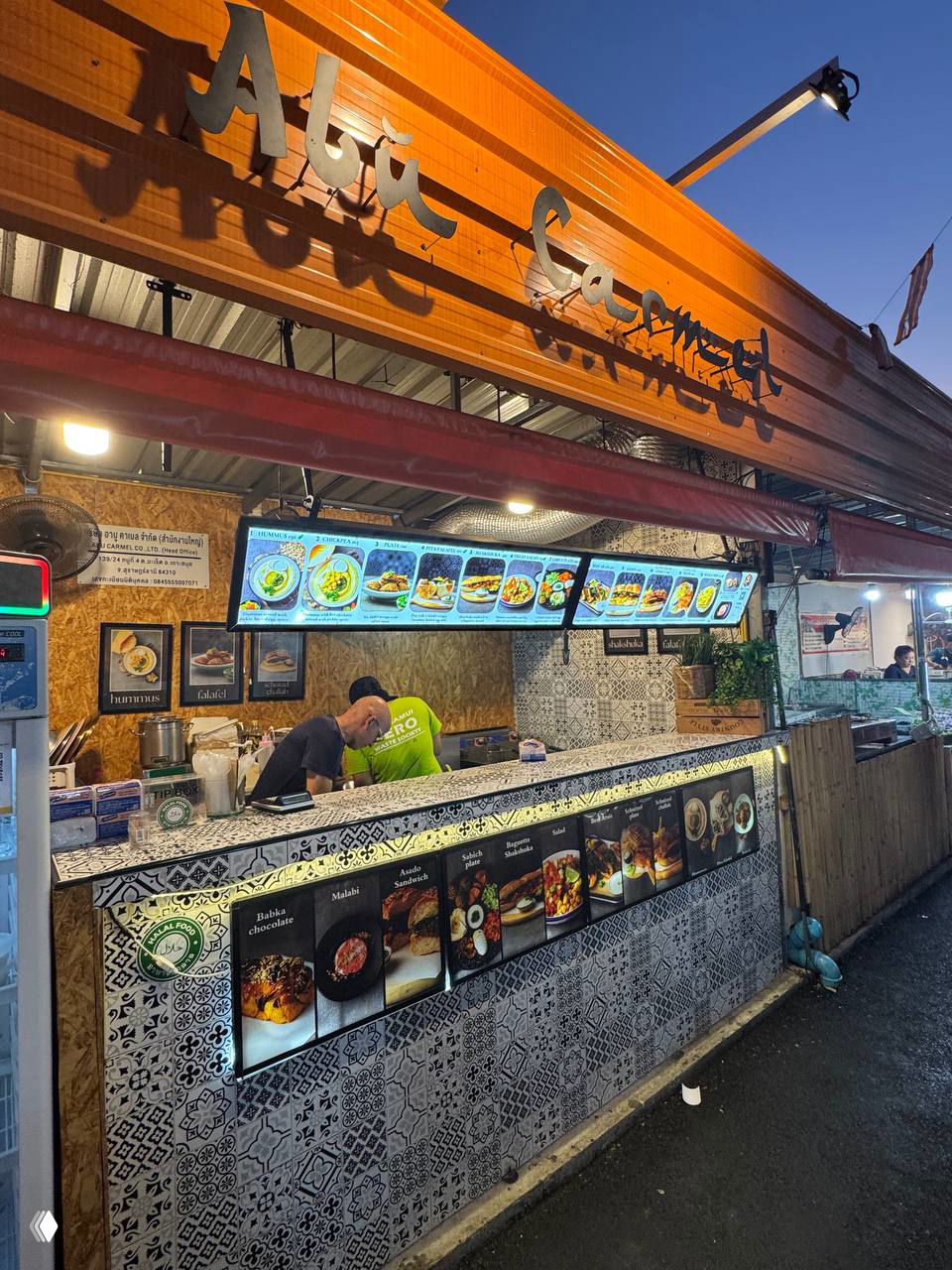 Фасад торговой точки Abu Carmen на ночном рынке Ламай вечером: вывеска, подсвеченное меню и прилавок с готовкой блюд.