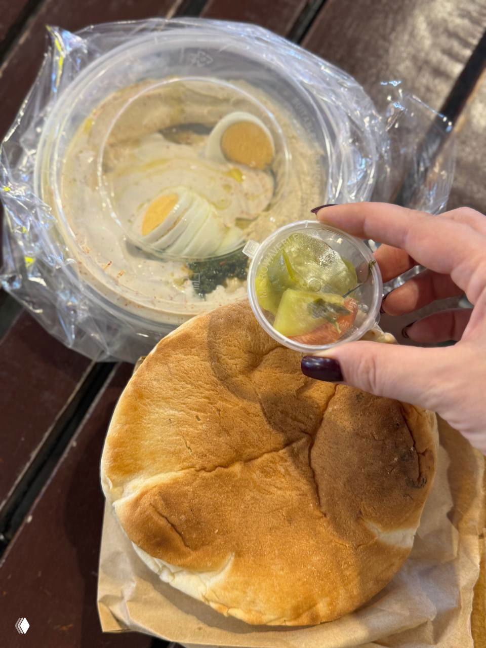 Плоская горячая пита, в руках маленькая банка с соусом/маринадом на фоне контейнера с хуммусом — вид с рук в кадре.