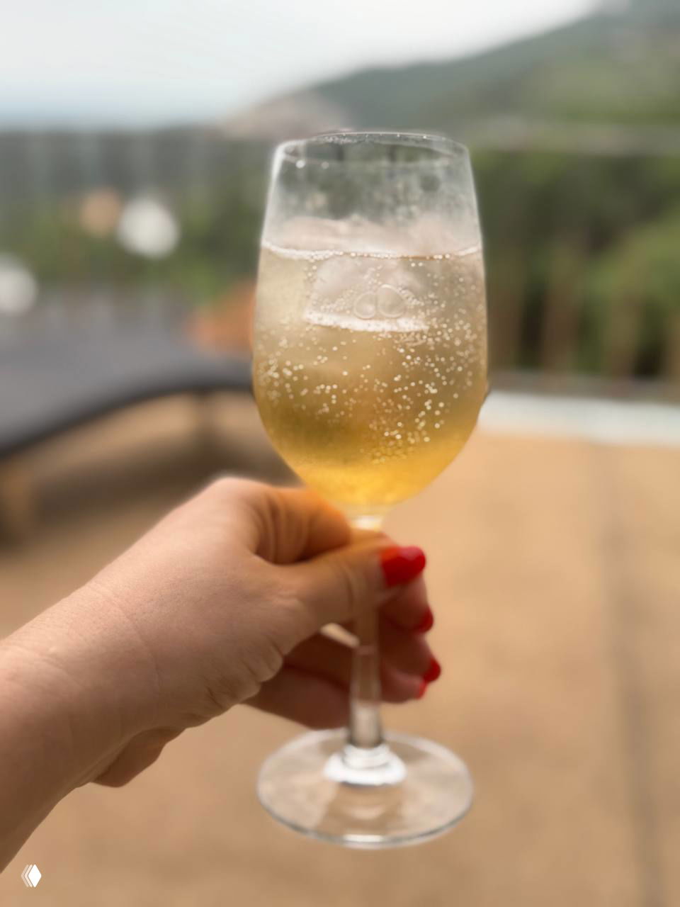 Бокал с игристым тайским умешу и льдом на фоне балкона и размытых холмов — золотистый пузырящийся напиток.