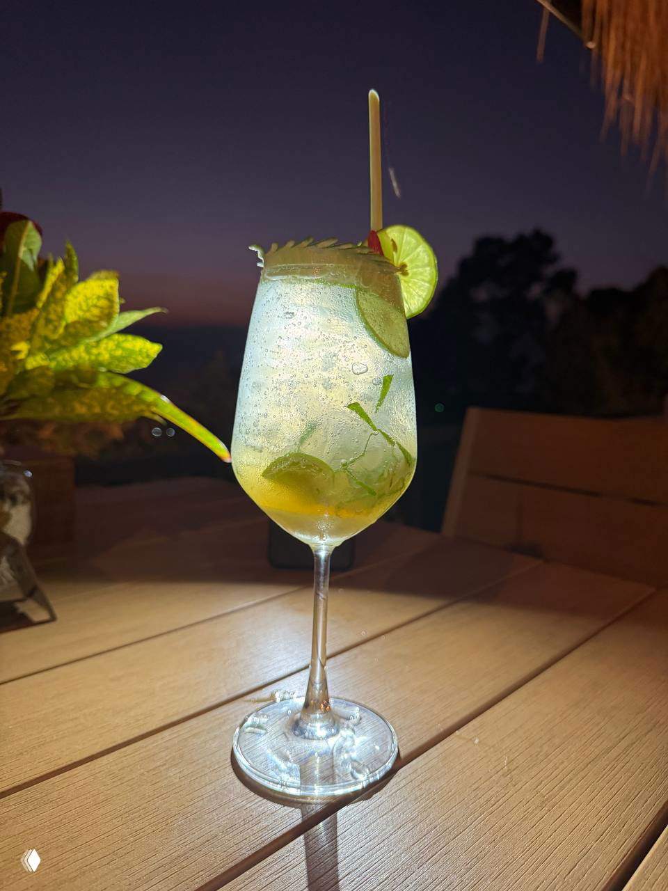Бокал с прозрачным коктейлем, лаймом и льдом на деревянном столе в сумерках — фотография коктейля Coconut Clouds или похожего напитка.