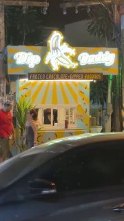 Ночью освещённый фуд‑ларёк Dip Daddy с неоновым логотипом и изображением банана, вид с улицы, вокруг проезжает машина — общая уличная сцена.