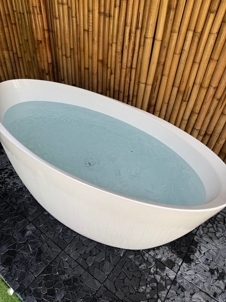 Белая овальная ванна со светлой холодной водой или льдом на полу у бамбуковой стены — ванна со льдом в сауне Vihara.