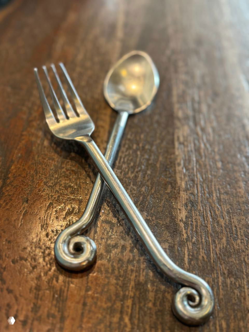 Детальный кадр: металлическая вилка и ложка с завитками, положенные крест-накрест на тёплой деревянной поверхности стола, демонстрируют стиль посуды.
