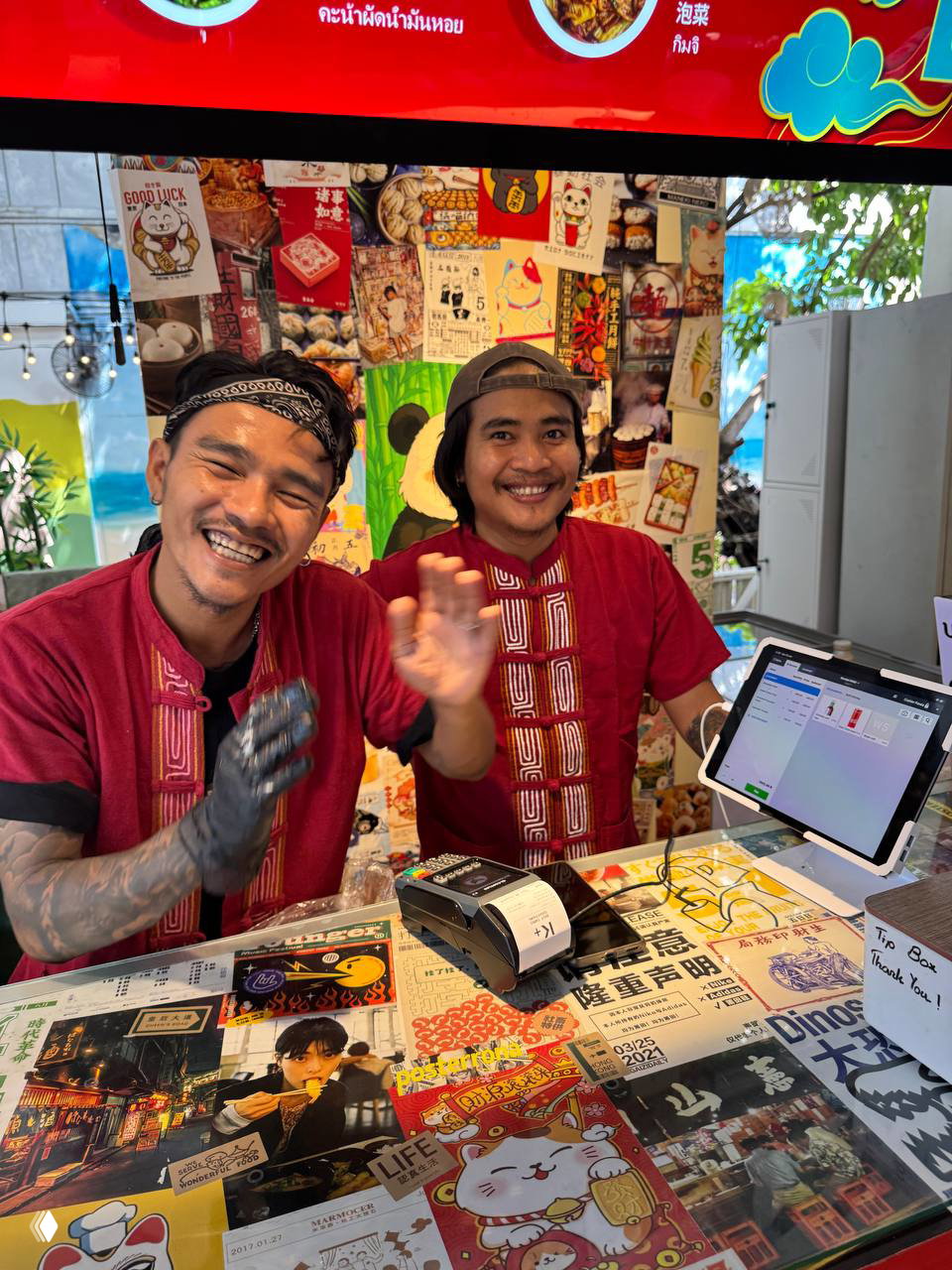 Два улыбающихся парня за стойкой в фуд-холе в красной униформе на фоне оформленного баннера с меню и китайскими декоративными элементами.