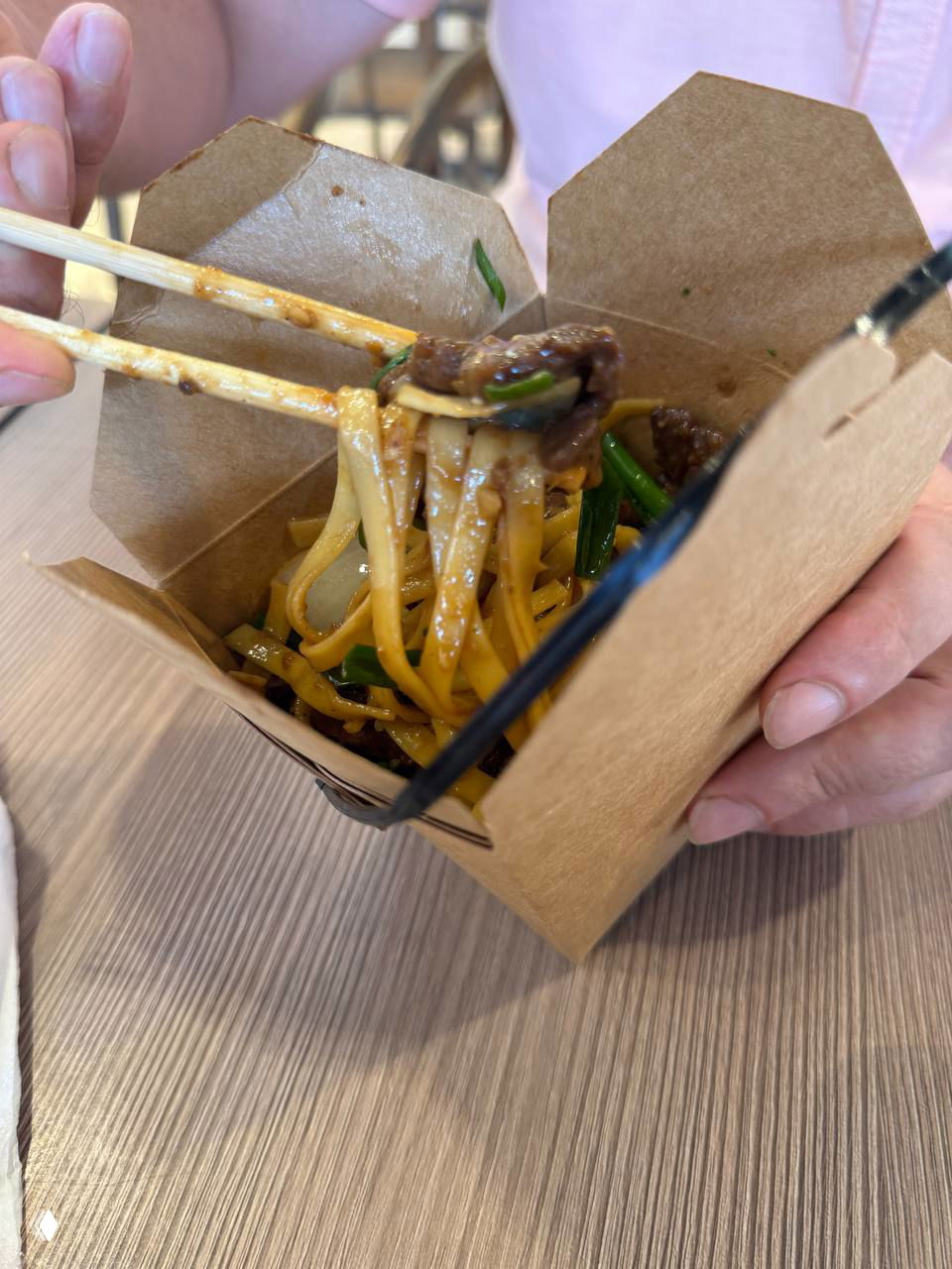 Крупный план лапши с кусочками говядины внутри коричневой одноразовой коробочки — видно соус, зелёные луковые стебли и текстуру мяса.
