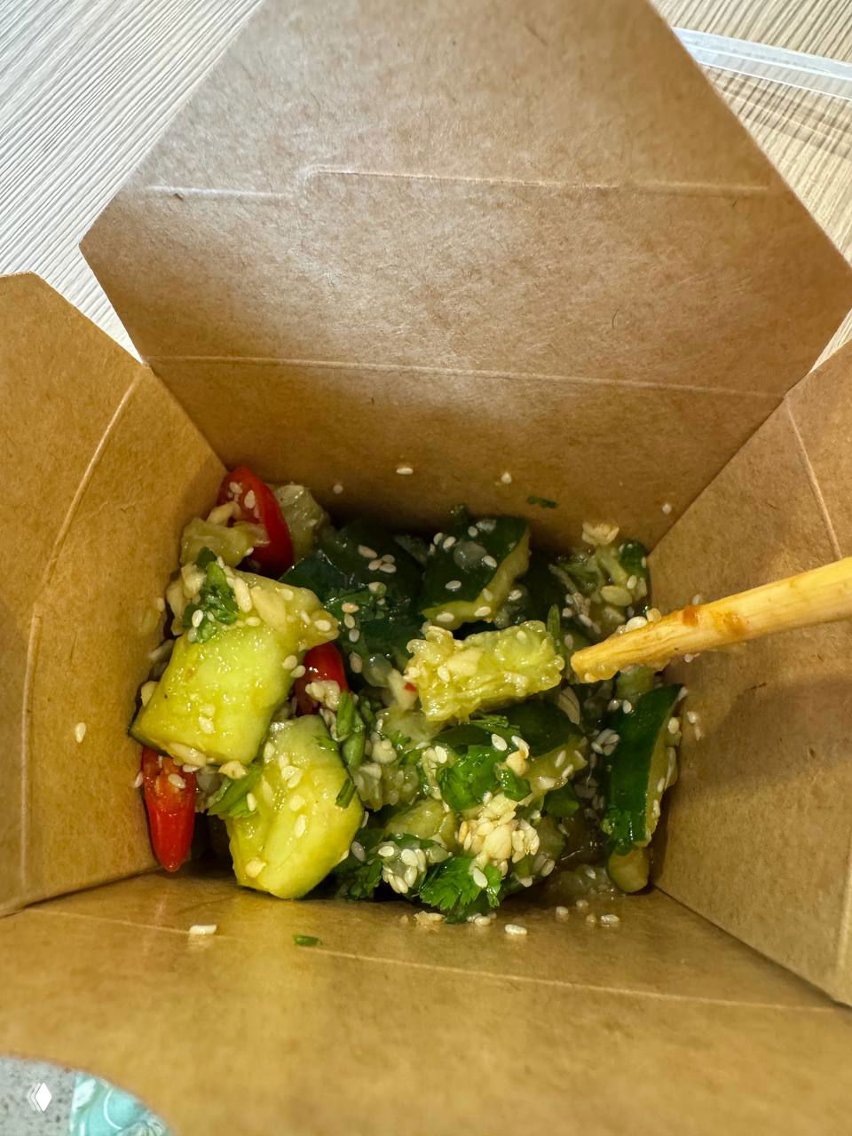 Салат из битых огурцов в одноразовой коробочке: кусочки огурца, кунжут, перец чили и зелень, всё смешано в лёгкой заправке.