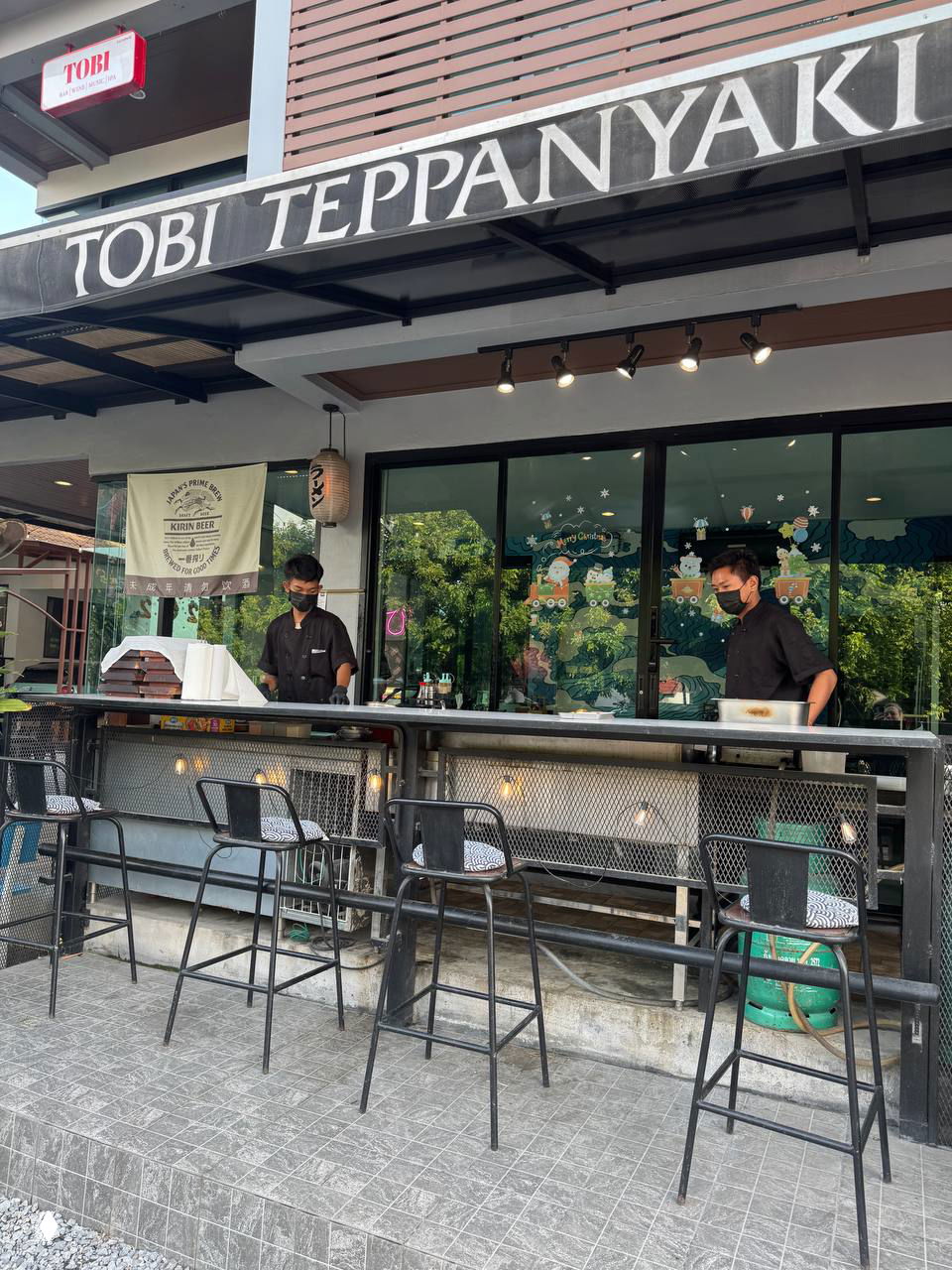 Фасад и стрит-бэнч Tobi Teppanyaki с открытой площадкой для жарки на теппане и двумя поварами в масках у металлической плиты.