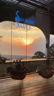 Вид на закат через стеклянную стену с вывеской Sora Above Samui: терраса, море и солнце на горизонте — атмосферное место для захода солнца.