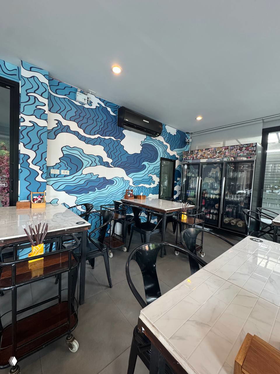 Интерьер заведения с японским муральным рисунком на стене, простыми столами и стульями в стиле столовой, видна холодильная витрина с напитками.