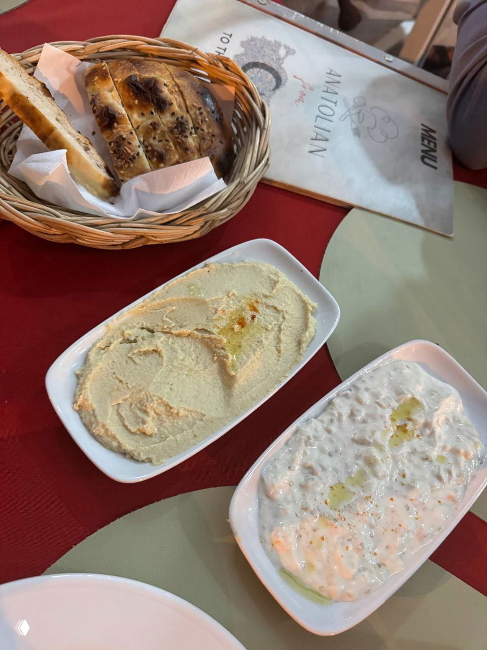 Блюда-закуски на столе: белые тарелки с хуммусом и йогуртовой закуской рядом с корзиной с хлебом и меню Anatolian.