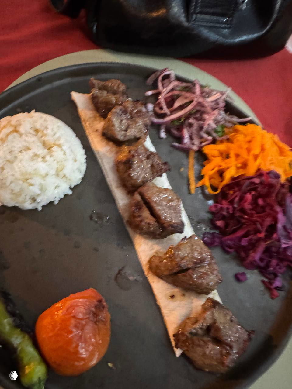 Порция кусков мяса кебаб на тонкой лепешке с рисом и тремя видами овощных гарниров — фото тёмнее, мясо выглядит сухим на тарелке.