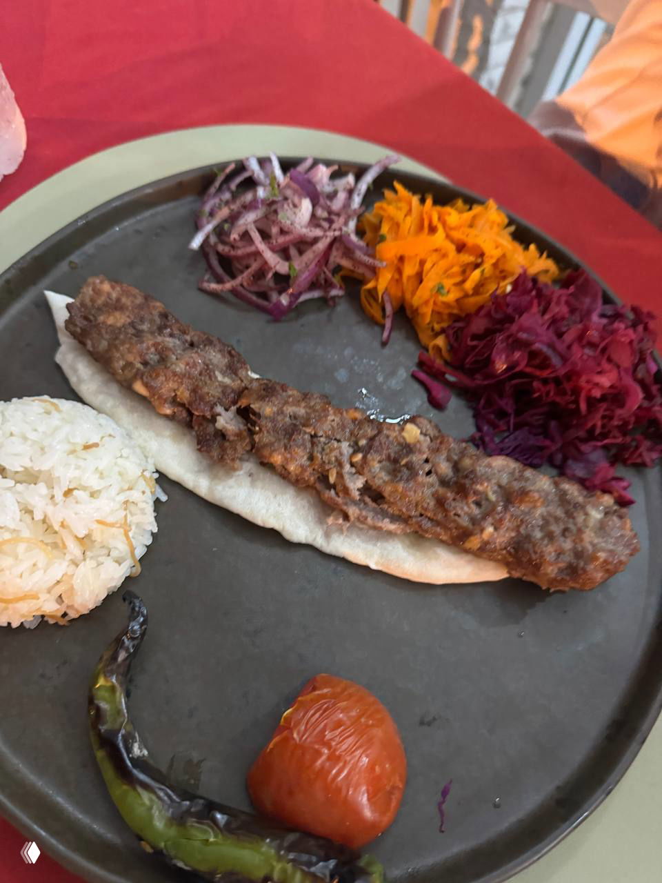 Длинный кебаб/люля-кебаб на лепёшке с жареным томатом и перцем, рядом порция риса и натёртые овощи, сервировка на чёрной тарелке.