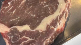 Крупный план сырого куска рибай сухой выдержки на металлической поверхности, видна мраморность мяса и жировая прослойка слева.