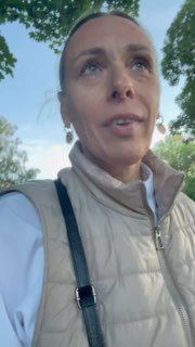 Кадр с автором канала на улице: верхняя часть тела в жилете, сумка на плече, на заднем плане зелёные кроны деревьев и небо.