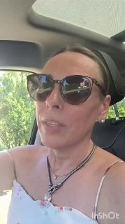 Крупный план женщины в солнцезащитных очках и летнем сарафане в салоне автомобиля; на фоне видны зелёные деревья и дневной свет через окно