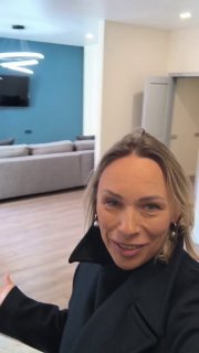 Ольга в квартире показывает интерьер: портрет агента на фоне гостиной с диваном, телевизором и современным потолочным светильником при показе объекта.