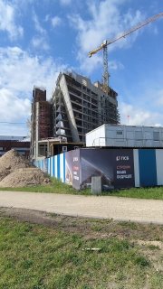 Панорамный кадр стройплощадки ЖК Триарт: частично возведённый корпус, башенный кран, кучи стройматериалов, вагончики и ограждение.