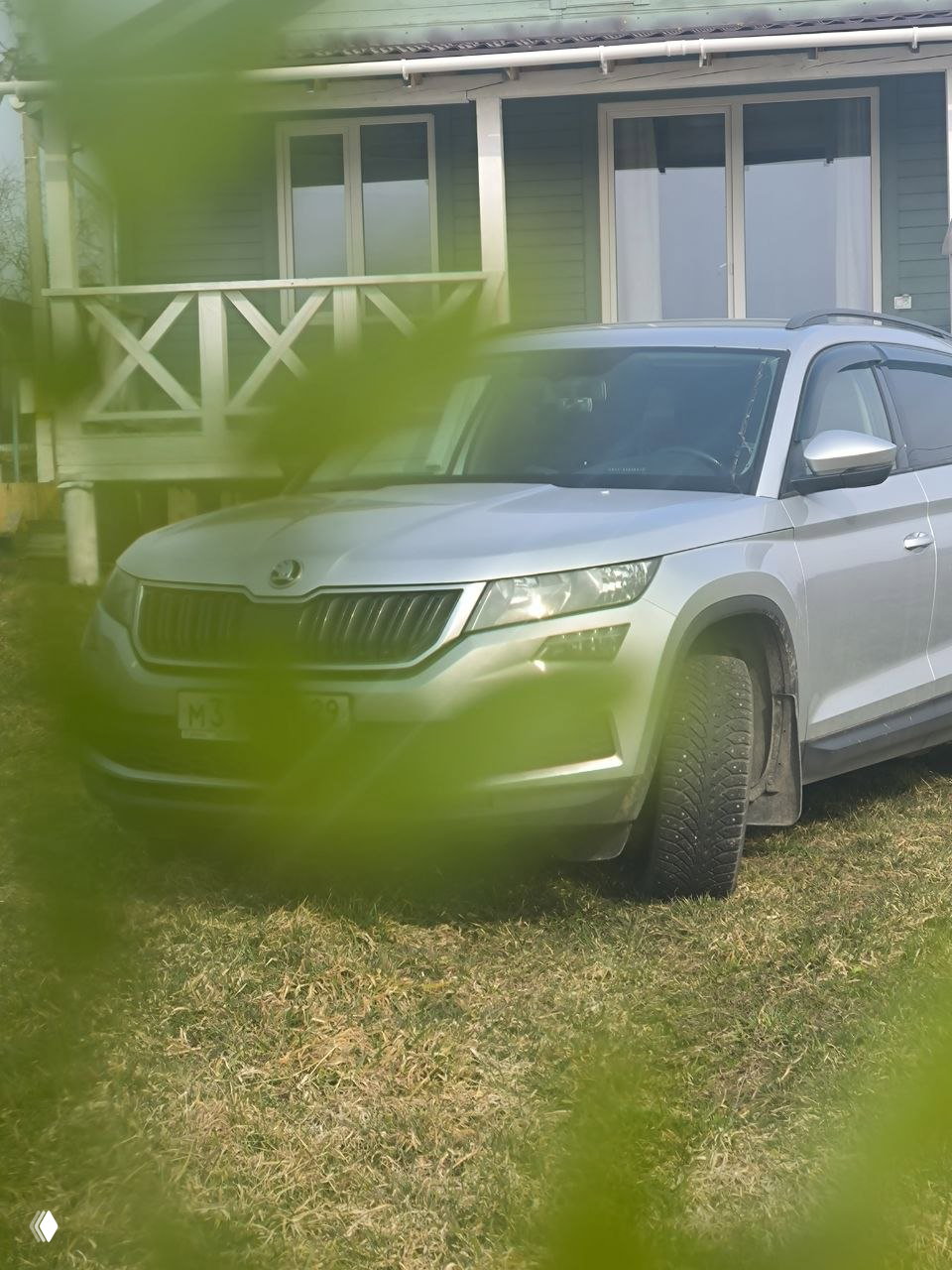 Серебристый автомобиль припаркован на газоне перед дачным домом, на переднем плане размытые зелёные листья и общий вид усадьбы