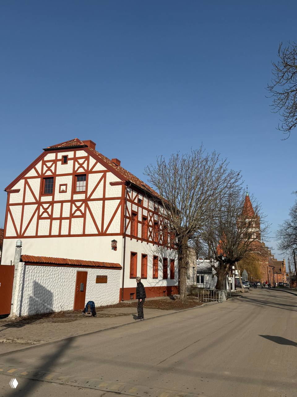 Фахверковый дом у широкой улицы в Правдинске, деревянные декоративные элементы и на заднем плане видна кирпичная башня кирхи и голубое небо.