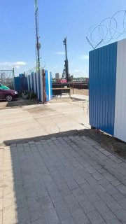 Вход на стройплощадку ЖК Триарт через ворота: вид на проход, плиточный тротуар, металлическое ограждение и техника за воротами.