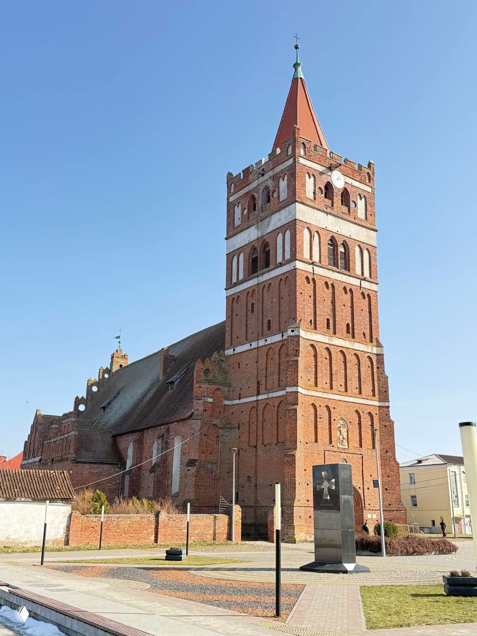 Вид на стройную кирпичную башню кирхи и прилегающую территорию с памятником на переднем плане; солнечный ясный день и чистое небо.