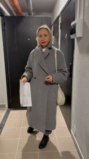 Женщина в сером пальто стоит в подъездном коридоре у входной двери кладовки, держит пакет и документы; видна плитка и щиток на стене.